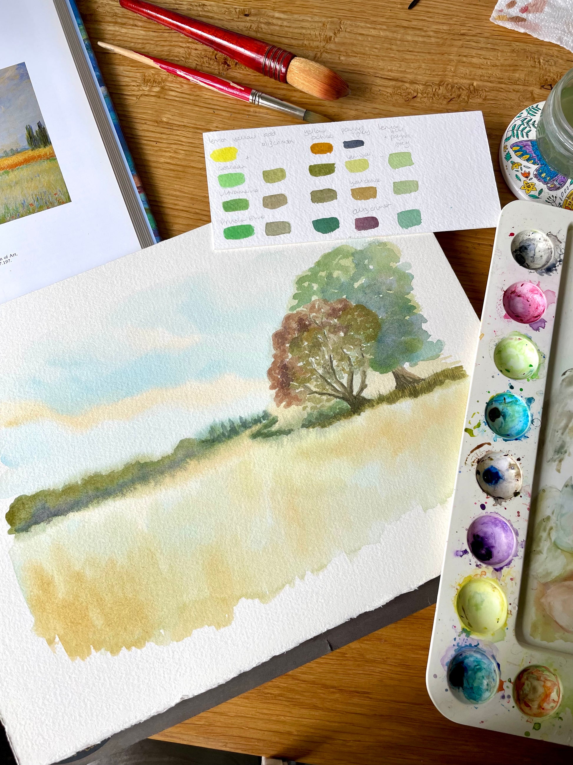 A watercolor landscape painting on textured paper, displayed on a wooden table alongside an artist's palette, paint tubes, brushes, and a color swatch card.
