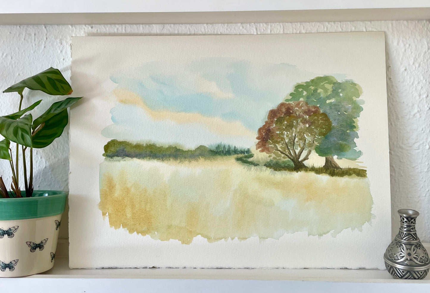 A watercolor landscape painting on textured paper, displayed on a shelf next to a potted plant and a small decorative vase. The painting depicts a tree in a grassy field under a soft blue sky.